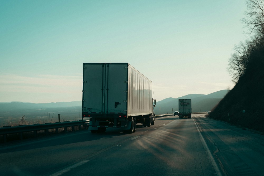 Dwie ciężarówki z naczepami typu chłodnia poruszające się po autostradzie w kierunku gór – ilustracja liniowego transportu międzynarodowego i optymalizacji tras.